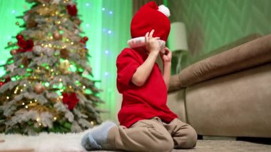 Bebek kanepenin yanında diz çökmüş, elinde Noel Baba şapkasıyla. Çocuk ayağa kalkar ve kaçar. Düşük açı. Arkaplanda Noel ağacı.