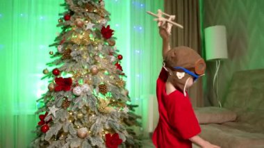 Evdeki Noel ağacının yanında pilot şapkası takan ve güneş gözlüğü takan şirin bir bebek oynuyor. Küçük çocuk tahta uçakla oynuyor..