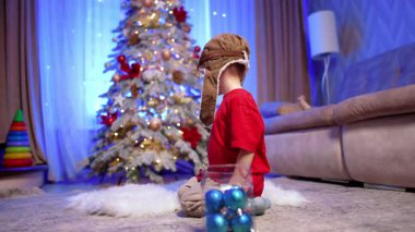 Havacı şapkalı sevimli bebek yerde oturuyor. Küçük çocuk Noel ağacı süslemeleriyle oynuyor..