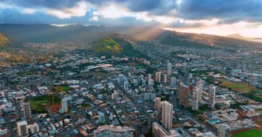 Batan güneş ışınları dağları aydınlatan gri bulutların arasından geçer. Honolulu, Hawaii, ABD 'nin yoğun şehir manzarasının üzerinde insansız hava aracı uçuşu..