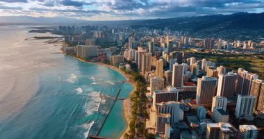 Waikiki tatil köyünün şehir manzarası batan güneşle aydınlanıyor. Okyanus dalgaları kumlu sahillere yuvarlanıyor. Arkaplandaki karanlık dağlar gri bulutların altında. Hava görünümü.