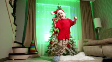 Noel Baba şapkalı mutlu çocuk odaya atlıyor. Tatlı çocuk Noel ağacının yanında eğleniyor..