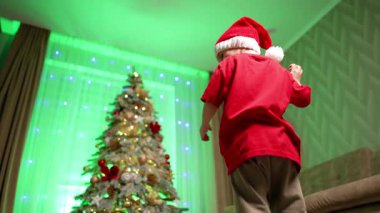 Noel Baba şapkalı sevimli çocuk kameranın yanına döndü. Noel ağacının yanında dans eden mutlu çocuk. Düşük açı görünümü.