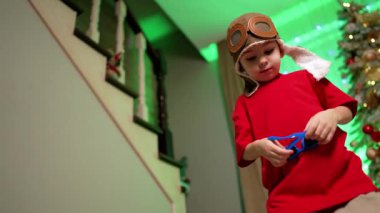 Havacı şapkalı ve kırmızı tişörtlü bebek Noel ağacında duruyor. Çocuk güneş gözlüğü takıyor ve uzaklaşıyor. Düşük açı görünümü.