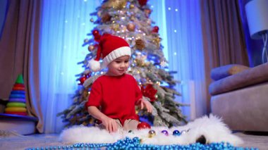 Noel Baba şapkalı şirin, mutlu çocuk Noel ağacının yanındaki yumuşak ekose üzerinde oturuyor. Sevimli çocuk oyuncak dekorasyonlarıyla oynuyor. Düşük açı görünümü.