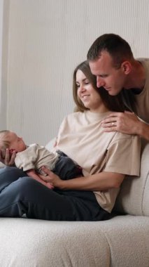 Uzun saçlı, beyaz bir kadın kanepeye oturup yeni doğan bebeğini kucağında tutuyor. Adam arkada duruyor ve omuzlarını okşuyor. Dikey video