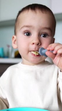 Sevimli erkek bebek kaşıktan yiyor. Güzel çocuk mutfakta oturuyor ve yemek yiyor. Kapatın. Dikey video