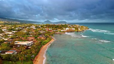 Bereketli yeşillikler arasında birden fazla evi olan güneşli bir sahil. Gökyüzünü kaplayan dramatik bulutlar. Honolulu, Hawaii, ABD. Hava görünümü.