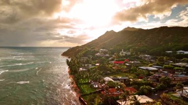 Dağ eteğindeki yeşil alanda bir sürü villa var. Bulutlu gökyüzündeki güneş manzarayı kamaştırır. Okyanus dalgaları kıyıya doğru yuvarlıyor. Honolulu, Hawaii, ABD. Hava görünümü.