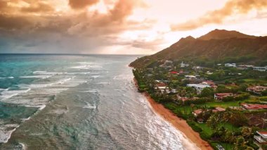 Okyanus dalgaları kumlu sahile ulaşır. Dağ eteğinin yeşil bölgesindeki yerleşim bölgesine hava perspektifi. Bulutlarda turuncu bulutlar. Honolulu, Hawaii, ABD.