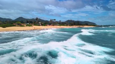 Güzel köpüklü dalgalar Hawaii kıyılarına vuruyor. Kumsal ve dağların siluetleri arka planda.