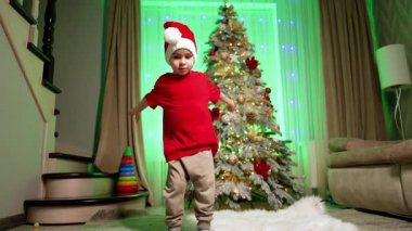 Noel Baba şapkası, kırmızı tişört ve bej pantolon giyen beyaz çocuk Noel ağacının yanında dans ediyor. Sevimli çocuk tatillerde eğleniyor..