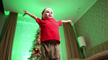 Kırmızı tişörtlü ve Noel Baba şapkalı yakışıklı çocuk zıplar, arkasını döner ve kameranın yanında el sallar. Düşük açı. Çocuklar Noel ağacında dans eder..