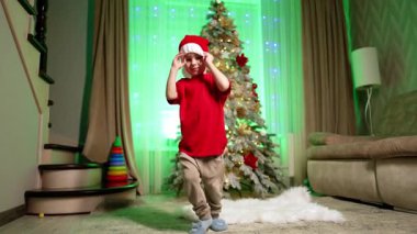 Noel Baba şapkalı aktif çocuk odada dans ediyor. Mutlu enerjik çocuk Noel ağacının yanında zıplıyor..