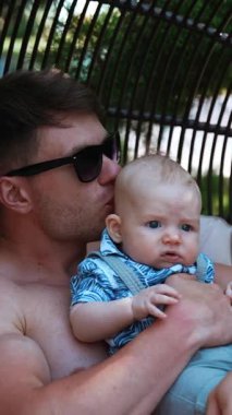 Babası küçük oğlunu kucağına aldı ve onu başından öptü. Baba, bebeğiyle dışarıda dinleniyor, dikey video.