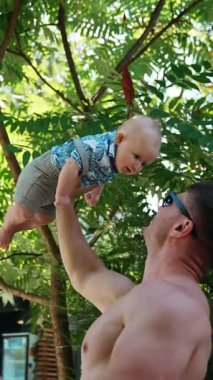 Kafkasyalı, gövdesi çıplak, bir elinde bebeği kafasının üstünde tutan güçlü bir adam. Baba küçük bir çocukla oynuyor, dikey video.