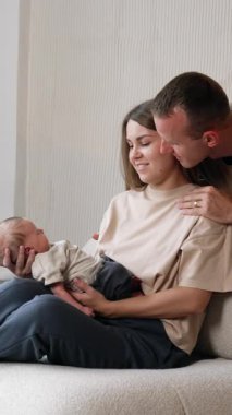 Genç bir kadın, elinde yeni doğmuş bir bebekle kanepede oturuyor. Sevgi dolu koca ve baba kanepenin arkasında dikey videonun arkasına yaslandı.