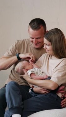 Yeni doğmuş bir bebeği olan beyaz bir aile. Ebeveynler kanepeye oturup annelerinin ellerinde bebeklerine bakarlar. Baba çocuğun elini dikey videoya çeker.