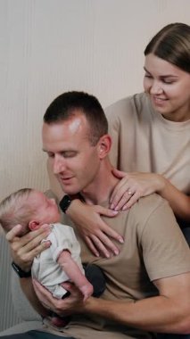 Yeni doğmuş baba, yeni doğan oğluna hayranlıkla bakıyor ve onu elinde tutuyor. Annem kocasının arkasında dikey videoda oturuyor.