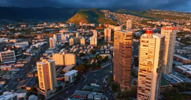 Batan güneşin son ışınları Honolulu 'nun yüksek binalarının tepelerini ve dağ yamacındaki evleri aydınlatıyor. Kara yağmurlu bulutlu manzara gökyüzünü kaplıyor. Hava perspektifi.