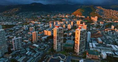 Batan güneşle aydınlatılmış benzer iki gökdelene yakın uçuyor. Arkaplandaki kayalar kara bulutlarla kaplıdır. Honolulu, Hawaii, ABD.