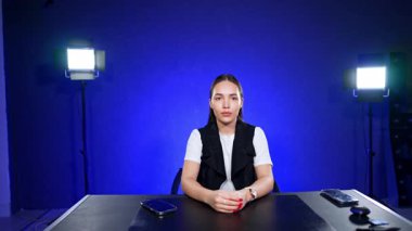 Beyaz kız mavi arka planda ciddi bir yüz ifadesiyle masada oturuyor. Blogcunun arkasında ışıklar yanıyor. Stüdyoda çekim.