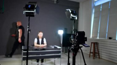 Kadın blogcu stüdyoda masada oturuyor. Kameraman etrafta dolaşıp ışık ve ekipmanları kontrol ediyor..