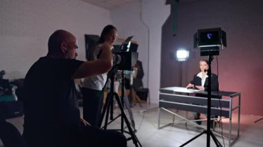 Video ekibi fotoğraf stüdyosunda çalışıyor ve blog içeriği oluşturuyor. Masada oturan kadın blogcu kamerayla konuşuyor. Fotoğraf çeken adam..