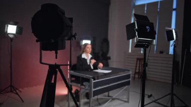 Profesyonel aydınlatma ekipmanları stüdyoda masa başında oturan bir kadının etrafında kurulur. Blogcu kamerayla konuşur ve elleriyle hareket eder.