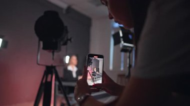 Beyaz tişörtlü uzun saçlı kız akıllı telefondan video çekiyor. Blogcu masasında oturup blogu için konuşuyor.