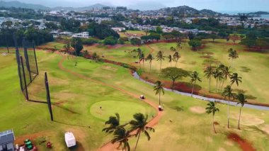 Palmiye ağaçlarının yetiştiği golf sahası. Bazı oyuncular sahada. Arka planda alçak katlı bir yerleşim alanı. Oahu, Hawaii, ABD. Hava görünümü.