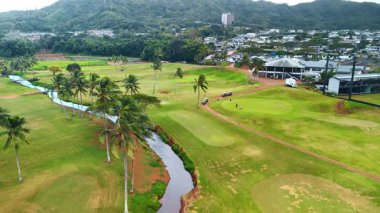 Üzerinde nehir ve palmiye ağaçları olan büyük yeşil bir golf sahası. Çok az erkek oyun oynar ve golf arabalarına kadar yürür. Hava görünümü.