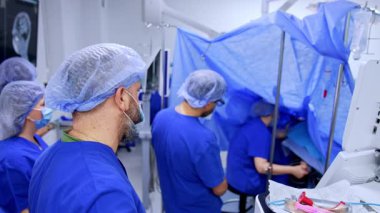 Mavi üniforma giyen bir grup doktor beyin cerrahisini inceliyor. Ameliyat yapan cerrah ameliyat alanında mavi çarşafla kaplanmış olarak oturuyor..