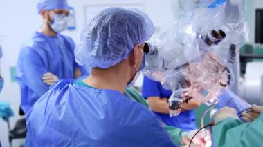 Şapkalı ve bornozlu erkek cerrahın mikroskoba bakışları. Tanınmayan asistan doktor için aleti tutuyor. Sıhhiye arka planda duruyor.