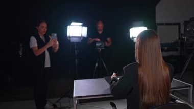 Esmer, uzun saçlı, sırtüstü uzanmış, yüzü çoraplara dönük bir şekilde kameraya bakıyor. Arka plandaki kız telefonuyla video çekiyor ve kameraman profesyonel kamera kullanıyor. Stüdyo günlüğü görüntüleri.