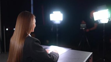 Uzun düz saçlı, pozitif esmer kadın masanın arkasında kameraya yarım yamalak oturuyor. Soffits 'in ışıkları blogcunun video kaydını aydınlatıyor. Arkaplanda erkek fotoğrafçı.