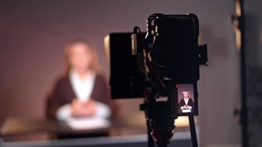 Tripodun üstündeki profesyonel kamera bir kadın blogcuyu çekiyor. Uzun saçlı bayan konuşuyor..