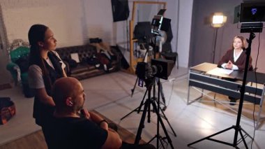 Kadın ve erkek sahne arkası çalışanları masada oturan blogcuya bakar. Modern ekipmanlar esmer bayanın videosunu çekiyor. İçerik oluşturma günlüğü.