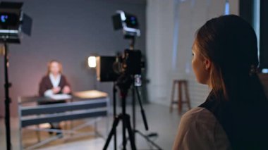 Kameranın yanında duran genç bayanın dikiz görüntüsü. Masadaki tanınmayan kadın blog içeriği için kameraya konuşuyor. Bulanık arkaplan.