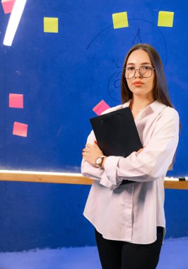 Not defteriyle kendine güvenen genç bir profesyonel. Açık renk tişört giymiş odaklanmış genç bir kadın renkli bir duvarın önünde elinde bir defter tutarak kameraya bakıyor..