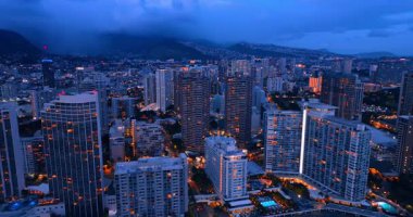 Modern şehirde alacakaranlık vakti. Çok katlı apartman blokları ışıkları açıyor. Kalın bulutlar arka plandaki dağları kaplıyor. Honolulu, Hawaii, ABD. Hava görünümü.
