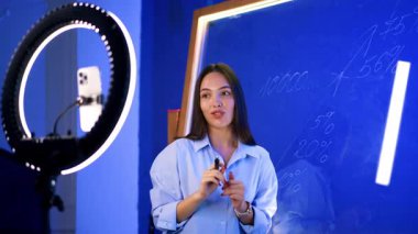 Gömlek giyen esmer, kameraya konuşan cam panoya numaralar gösteren bir kadın. Kadın blogcu telefonla video çekiyor.