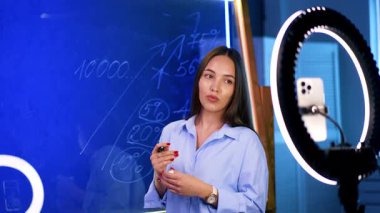 Kadın cam tahtada sayıları açıklayan ve tasvir eden kameralarla konuşuyor. Blog konsepti. Telefon kamerasında halka ışığıyla görüntüler.