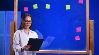 Gözlüklü, beyaz gömlekli, elinde dosya olan çekici beyaz bir kadın. Cam tahtanın yanında kendine güvenen bir iş kadınının portresi..