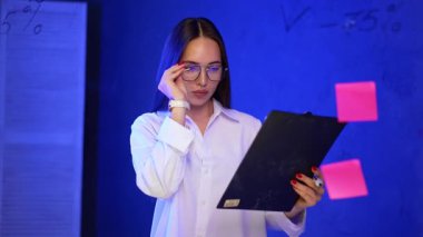 Beyaz bluzlu güzel esmer gözlük takıyor ve cam bir tahtanın yanına geliyor. İş kadını siyah kalemle rakamlar yazıyor..