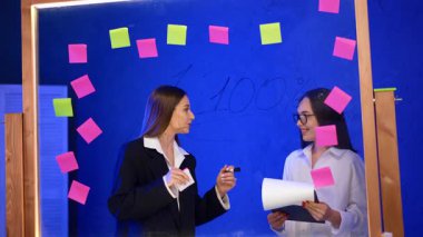 Cam tahtanın arkasında üzerinde çıkartmalar olan iki bayan var. Siyah ceketli bir kadın tutkuyla konuşuyor ve el kol hareketi yapıyor..