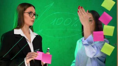 İş kadınları üzerinde çıkartmalar olan cam tahtaya bakarlar. İletişim, işbirliği ve inşa stratejileri.