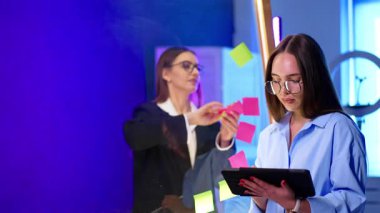 Siyah ceketli esmer, cam levhadan çıkartmalar çıkarıyor. Önplandaki kadın tablet kullanıyor.