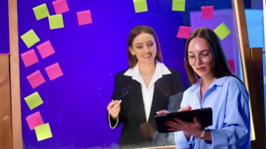 Cam tahtanın arkasında durup tablete bakan iki bayan meslektaşım var. Kadınlar video içeriği konuşmalarını izliyor.