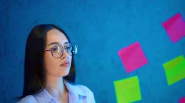 Koyu renk saçlı, gözlüklü ve tişörtlü bir kadın cam tahtaya etiket yapıştırıyor. İş konsepti.